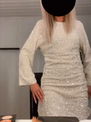 Paljettklänning i ljus champagne - Snygg kort klänning täckt av glittrande paljetter i en ljus champagnefärg. Klänningen har lång ärm och rund halsringning, perfekt för dig som vill sticka ut. Materialet känns mjukt och skimrande, och modellen är rak och enkel. (Den är sydd längst ner på höger sida, bild 6)