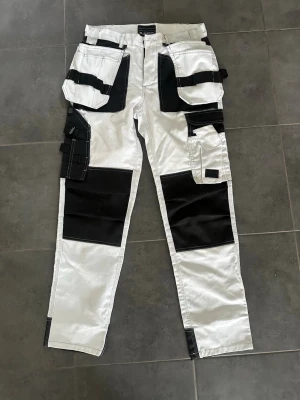 Arbetsbyxor från Bluewear, vit/svart - Säljer ett par vita arbetsbyxor från Bluewear med svarta förstärkningar och många praktiska fickor. Byxorna har flera detaljer för verktyg och är perfekta för dig som gillar funktionella plagg med cool look.