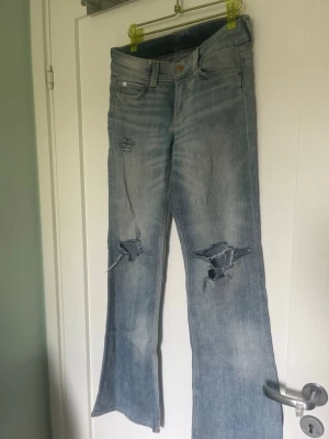 Ljusblå slitna bootcut jeans - Säljer ett par ljusblå jeans med bootcut-modell och gjort egna hål  Jeansen har klassisk femficksdesign, låg midja och är tillverkade i denim. Perfekta för en avslappnad och trendig look.