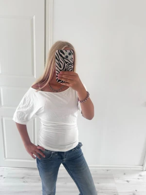 Vit offshoulder topp - Säljer en vit offshoulder topp med korta ärmar och figurnära passform. Toppen har en bred halsringning som ger en snygg och avslappnad look. Perfekt att matcha med jeans eller kjol för en trendig outfit.