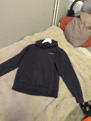 Svart hoodie från Peak Performance  - Svart hoodie från Peak Performance med liten vit logga på bröstet. Klassisk modell med huva och stor magficka framtill. Mjuk bomullsblandning och ribbade muddar vid ärmslut och nederkant. Perfekt för en chill och stilren look.