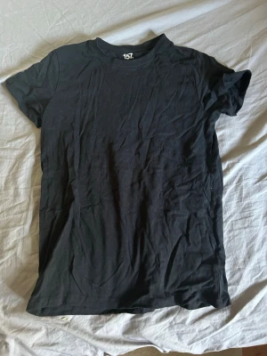 Svart basic t-shirt från 157 - En enkel svart t-shirt från 157 med rund hals och korta ärmar. Tillverkad i mjuk bomull och har en klassisk passform som funkar till allt. Perfekt basplagg för garderoben.
