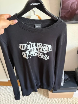 Svart långärmad tröja Just Cavalli - Svart långärmad tröja från Just Cavalli med ett vitt tryck framtill i en cool, trasig stil. Tröjan har rund halsringning och ribbade muddar vid ärmslut och nederkant. Perfekt för dig som gillar streetwear och vill sticka ut.