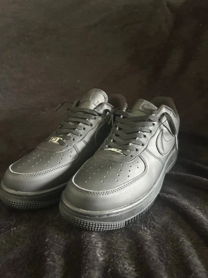 Svarta Nike Air Force 1 sneakers - Säljer ett par klassiska helsvarta Nike Air Force 1 sneakers i lågt utförande. Skorna har ovandel i slätt läder, perforeringar på tån för ventilation och den ikoniska swoosh-loggan på sidan. Tjock sula och svarta skosnören ger en clean look.