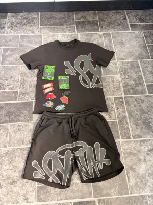 Svart syna world set med graffiti-tryck - Säljer ett svart set med t-shirt och shorts. Båda plaggen har stora gråa graffiti-inspirerade tryck. T-shirten har även färgglada patch-liknande tryck framtill. Shortsen har resårmidja med snörning och en bakficka. Materialet ser ut att vara mjukt och bekvämt, perfekt för en avslappnad stil.