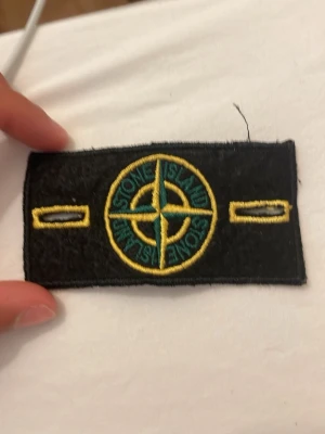 Stone Island tygmärke svart/gult - Säljer ett klassiskt Stone Island tygmärke i svart med gul och grön brodyr. Märket har den ikoniska kompassen i mitten och är rektangulärt med broderade knapphål på sidorna. Perfekt att sy eller fästa på jacka eller tröja.