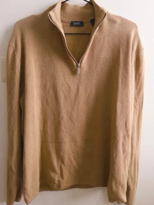 Beige stickad tröja med halv dragkedja - Beige stickad tröja från Esprit med halv dragkedja och hög krage. Tröjan har lång ärm och är tillverkad i ett mjukt, ribbat material som ger en avslappnad och stilren look. Perfekt för lager-på-lager och enkel att matcha med olika outfits.