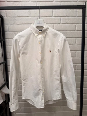 Vit skjorta med broderad logga - Klassisk vit skjorta i bomull med lång ärm och knappar framtill. Skjortan har en broderad logga på bröstet och en stilren krage. Perfekt för dig som gillar en clean och tidlös look.