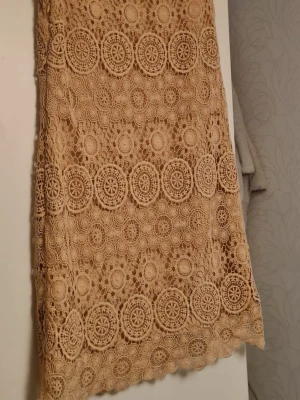 Beige spetsklänning med volangkant - Säljer en beige kortklänning i spets med vackert cirkelmönster och volangkant nedtill. Klänningen är ärmlös och har en rak passform som ger en avslappnad men ändå elegant look. Perfekt för sommarens alla tillfällen!