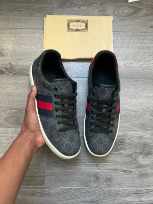 Gucci sneakers med monogram och röd detalj - Snygga svarta Gucci sneakers med klassiskt GG-monogram över hela skon och röda samt mörkblå ränder på sidorna. Skorna har svarta skosnören, vit sula och låg profil. Perfekta för dig som vill sticka ut med en lyxig och trendig look.
