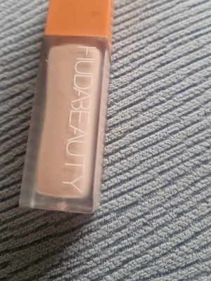 Huda Beauty flytande concealer - Flytande concealer från Huda Beauty i en rektangulär genomskinlig plastförpackning med orange lock. Färgen är ljusbeige och passar för att täcka mörka ringar eller ojämnheter. Enkel att applicera med applikator och ger ett naturligt resultat.obs den är använd några gånger 