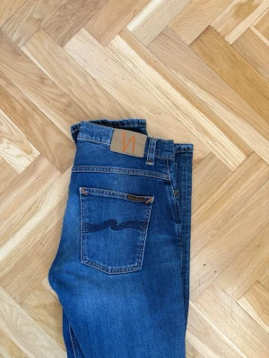 Blå jeans från Nudie Jeans - Säljer ett par klassiska blå jeans från Nudie Jeans med smal passform och snygga slitningar. Jeansen har fem fickor, orange sömmar och den ikoniska vågen på bakfickan. Perfekta för dig som gillar en tidlös och avslappnad stil.