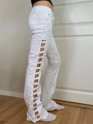 Vita jeans med cut-out detaljer - Säljer ett par vita jeans med unika cut-out detaljer längs sidorna. Jeansen har låg midja, klassiska fickor och är tillverkade i bomull. Perfekta för dig som vill sticka ut med en trendig och edgy stil.