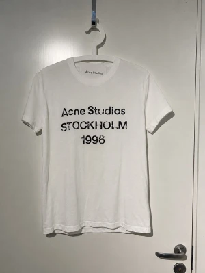 Vit Acne Studios t-shirt Stockholm - Vit t-shirt från Acne Studios med svart text 'Acne Studios STOCKHOLM 1996' på bröstet. Klassisk rund hals och korta ärmar. Tillverkad i mjuk bomull för en skön och avslappnad look.