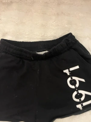 Svarta shorts från H&M med tryck - Svarta shorts från H&M med resår i midjan och ett vitt tryck med siffrorna '1991' på ena benet. Sköna och enkla att matcha, perfekta för chill dagar eller träning. Gjorda i mjukt bomullsmaterial.
