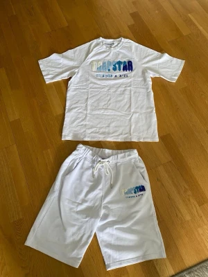Vitt set med tryck från Trapstar - Säljer ett vitt set från Trapstar med t-shirt och shorts. Båda delarna har färgglatt Trapstar-tryck i blått och vitt samt texten 'It's a secret'. T-shirten har rund hals och korta ärmar, shortsen har dragsko i midjan och sidofickor. Perfekt för en avslappnad stil.