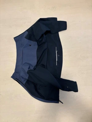 Mörkblå och blå vindjacka - Snygg vindjacka i mörkblått och blått med dragkedja och hög krage. Jackan har en minimalistisk design med diskreta detaljer och är tillverkad i ett lätt material som passar perfekt för blåsiga dagar. Fodret är svart och jackan har praktiska fickor. Jackan är lite skitig men kan tvättas innan leverans! 