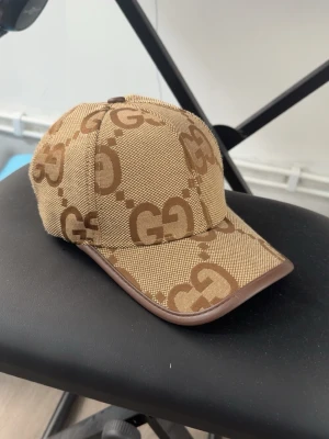 Beige keps med GG-mönster - Snygg beige keps med brunt GG-mönster och bruna detaljer längs skärmen. Kepsen har en klassisk form och är tillverkad i ett tåligt tygmaterial. Perfekt för dig som vill ha en trendig accessoar med streetwear-vibe.