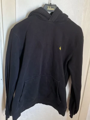 Lyle and Scott svart hoodie  - Säljer en svart hoodie med huva och känguruficka framtill. På bröstet finns en liten gul broderad logga. Hoodien har långa ärmar och ribbade muddar. Perfekt för en chill och avslappnad stil. Synlig blekning i tyget men går att få bort 
