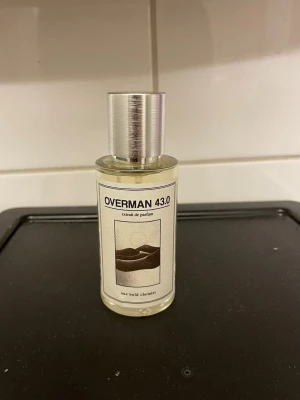 Overman 43.0 Extrait de Parfum - Inspirerad av Tom Ford Ombre Leather. En stilren parfymflaska med metallock och transparent glas, innehållande Overman 43.0 extrait de parfum. Flaskan har en minimalistisk etikett med konstnärlig design och texten 'one bold chemist'. Perfekt för dig som gillar unika dofter.