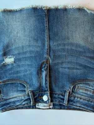Kort jeanskjol med fransad kant storlek 134/ XXXS från Zara  - Säljer en blå kort jeanskjol med fransad kant och slitna detaljer. Kjolen har klassiska fickor och bälteshällor, samt en knapp framtill. Perfekt skick! Storlek 134/ XXXS från Zara 