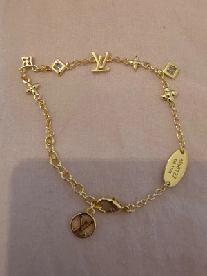 Louis Vuitton armband - Snyggt guldigt armband från Louis Vuitton med ikoniska LV-detaljer och små berlocker. Kedjan har flera olika former och en oval bricka med ingraverad kod. Perfekt accessoar för dig som gillar exklusiv och trendig stil.