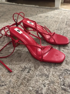 Röda strappy sandaler med klack - Snygga röda sandaler med tunna remmar och fyrkantig tå. Klackarna är medelhöga och sandalerna har långa snören som kan lindas runt ankeln. Tillverkade i ett glansigt material som ger en trendig look. Perfekta för dig som vill sticka ut.