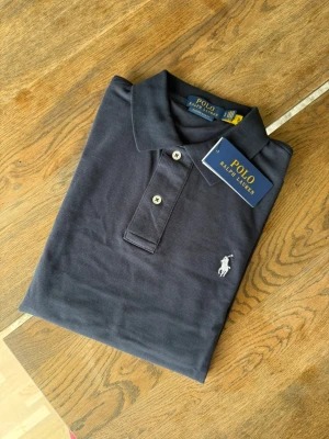 Mörkblå pikétröja från Polo Ralph Lauren - Klassisk mörkblå pikétröja från Polo Ralph Lauren med vit broderad logga på bröstet. Tröjan har krage, två knappar framtill och korta ärmar. Perfekt för en clean och stilren look.