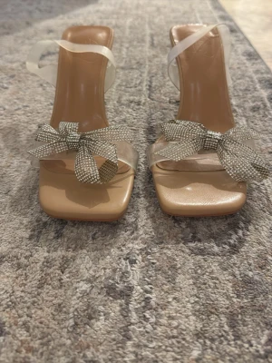 Beiga pumps med transparent rem och rosett - Snygga pumps med beige bas, transparenta remmar och fyrkantig tå. Dekorerade med glittrande silverrosetter framtill. Klacken är medelhög och smal, vilket ger en elegant siluett. Perfekta för att lyfta din outfit med en trendig touch.
