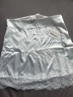 Vit satinkjol med spetsdetaljer - Snygg vit spetskjol i satin. Materialet är glansigt och mjukt, vilket ger en lyxig känsla. Helt ny och aldrig använd. 
