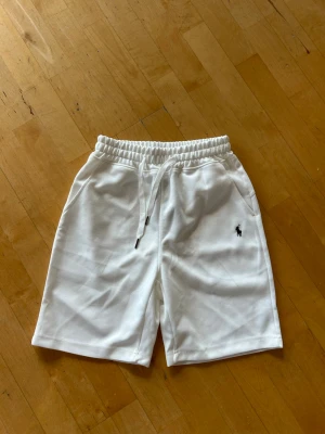 Vita shorts från Ralph Lauren med snörning - Snygga vita shorts från Ralph Lauren med elastisk midja och snörning. Shortsen har två sidofickor och en liten broderad Ralph Lauren logga på vänster ben. Tillverkade i mjukt material som känns skönt mot huden. Perfekta för varma dagar.