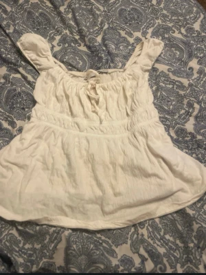 Vit blus med knyt i halsen - Söt vit blus med korta ärmar och knyt i halsen. Blusen har en luftig och avslappnad passform med lätt rynkade detaljer och är tillverkad i ett mjukt bomullsliknande material. Perfekt för varma dagar och enkel att styla med jeans eller kjol.