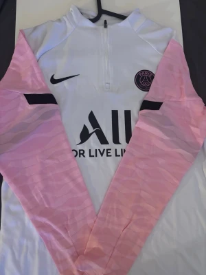 PSG träningströja med rosa ärmar - Säljer en snygg PSG träningströja från Nike med vita detaljer och rosa mönstrade ärmar. Tröjan har halv dragkedja, klubbmärke på bröstet och svart Nike-logga. Materialet är troligtvis polyester, perfekt för träning eller sport.