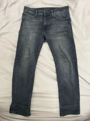 Grå Regular Clark jeans - Säljer ett par grå jeans med raka ben och klassisk femficksmodell. Jeansen har en snygg tvättad look med ljusare partier på låren och knäna. Tillverkade i ett mjukt denimtyg som är skönt att bära hela dagen. Size är 32/30