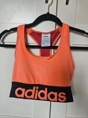 Orange sport-bh från adidas - Snygg sport-bh från adidas i orange med svart resår och stor logga framtill. Baksidan har racerback-design och meshpanel i rött för extra ventilation. Perfekt för träning och sportiga aktiviteter.