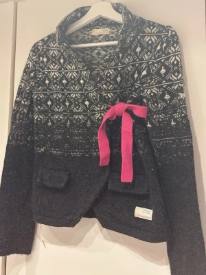 Mörkgrå kofta Odd Molly med mönster - Snygg mörkgrå kofta från Odd Molly med vitmönstrad överdel och en rosa knytband i midjan. Koftan har två fickor framtill och en unik etikett på sidan. Perfekt för dig som gillar stickade plagg med detaljer.