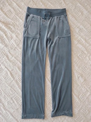 Juicy Couture velourbyxor ljusblå S - Snygga blåa mjukisbyxor i mjuk velour med bred resårmidja och dragsko. Byxorna har raka ben och två stora fickor framtill. Perfekta för chill dagar eller när du vill vara extra bekväm. Beninnerlängd ca 79 cm.