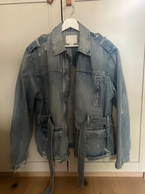 Oversized ljusblå jeansjacka med bälte - Säljer en oversized ljusblå jeansjacka med slitna detaljer, axelklaffar och bälte i midjan. Jackan har stora fickor framtill och råa kanter vid ärmslut och nederkant för en trendig, avslappnad look. Perfekt för dig som gillar streetstyle.