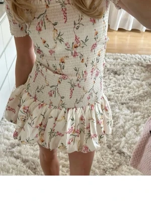 Kort blommig volangklänning - Supersöt kort klänning med blommigt mönster i rosa och grönt på vit botten. Klänningen har volang nedtill och korta ärmar. Perfekt för sommarens alla tillfällen och ger en romantisk vibe.