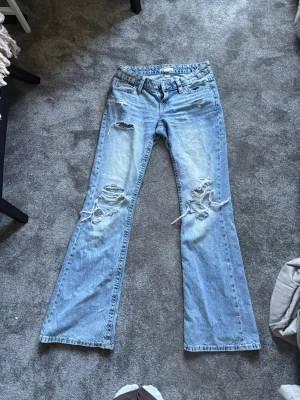 Lowrise Bootcut jeans med slitningar, ljusblå - Säljer ett par ljusblå bootcut jeans med slitningar och hål på både fram- och baksida. Jeansen har en lös passform och är tillverkade i klassisk denim. Perfekta för dig som gillar en avslappnad och trendig stil. Passar mig som normalt sätt har stl 32-36 i jeans och är 1,67m. Använda kanske 2 gånger.