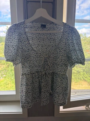 Blommig vit blus med puffärm - Söt blus i vitt med små blå blommor, puffärmar och knytband framtill. Blusen har v-ringning och smockdetaljer vid midjan för en feminin siluett. Perfekt till jeans eller kjol för en fräsch sommarlook.