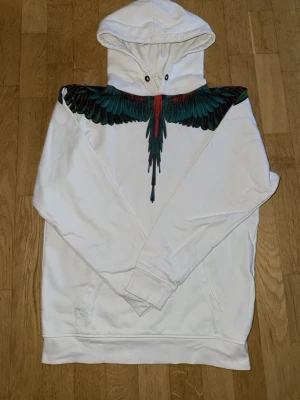 Marcelo burlon hoodie - Säljer en vit hoodie med stor huva och ett fett tryck av färgglada fågelfjädrar i svart, grönt och rött över axlar och bröst. Hoodien har känguruficka framtill och ribbade muddar. Perfekt för dig som vill sticka ut med en unik design. Det saknas snöre.