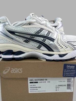 ASICS Gel-Kayano 14 vita sneakers - ASICS Gel-Kayano 14 sneakers i vitt med svarta och silverdetaljer. Skorna har en sportig design med meshpaneler och Ortholite-innersula för extra komfort. Snygga kontrasterande linjer och robust sula gör dem perfekta för en aktiv livsstil.