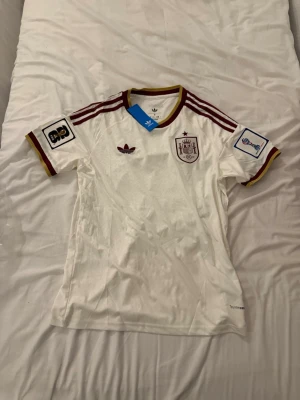 Vit Spanien Adidas retro matchtröja - Säljer en vit retro matchtröja från Adidas med Spaniens landslagsemblem på bröstet. Tröjan har vinröda och gula detaljer på krage och ärmslut samt tre klassiska Adidas-ränder på axlarna. Korta ärmar och broderade märken. Perfekt för fotbollsfans!