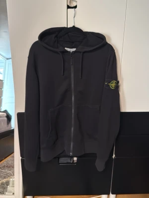 Svart zip hoodie från Stone Island - Säljer en svart hoodie med dragkedja från Stone Island. Klassisk huva med zip och två fickor framtill. Ikonisk Stone Island patch på vänster ärm. Perfekt för dig som gillar streetwear och vill ha en stilren look.