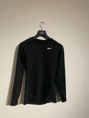 Svart långärmad träningströja Nike - Snygg svart långärmad träningströja från Nike med vit logga på bröstet. Tillverkad i ett stretchigt funktionsmaterial som andas, perfekt för träning eller löpning. Tight passform och rund halsringning. Nypris 1000kr 