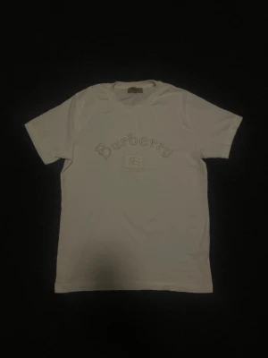 Vit Burberry t-shirt med broderi - Säljer en vit t-shirt från Burberry med snyggt broderat Burberry-tryck och emblem på bröstet. Klassisk passform och rund halsringning. Perfekt för dig som gillar stilrena och exklusiva plagg. Tröjan är i M men kan även passa L i olika fall.  🚨‼️pris kan diskuteras vid snabb affär‼️🚨