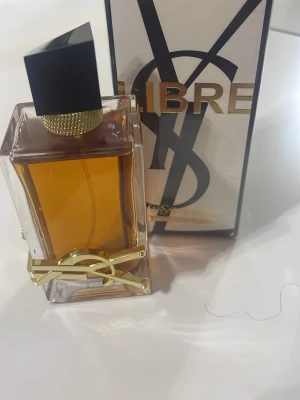 Yves Saint Laurent Libre parfym - Libre från Yves Saint Laurent är en lyxig parfym i en rektangulär glasflaska med guldiga detaljer och svart asymmetrisk kork. Flaskan har en varm bärnstensfärgad vätska och pryds av YSL-loggan i metall. Förpackningen är elegant med stora bokstäver och grafisk design.