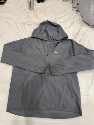 Grå vindjacka från Nike - Snygg grå vindjacka från Nike med dragkedja framtill och två sidofickor. Jackan har en klassisk huva och är tillverkad i ett lätt och smidigt material. Perfekt för blåsiga dagar och sportiga outfits. Nike-logga på bröstet ger en sportig look.