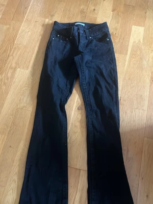 Svarta bootcut jeans med fickdetalj - Säljer ett par svarta bootcut jeans med snygga fickor bak som har lock och silverfärgad knapp. Jeansen har klassisk femficksmodell och är tillverkade i ett mjukt denimtyg. Perfekta för dig som gillar en retroinspirerad look.